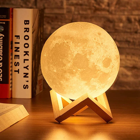 A glowing moon lamp on a wooden stand sits on a tabletop in a warm indoor setting, with books and a brick wall in the background.