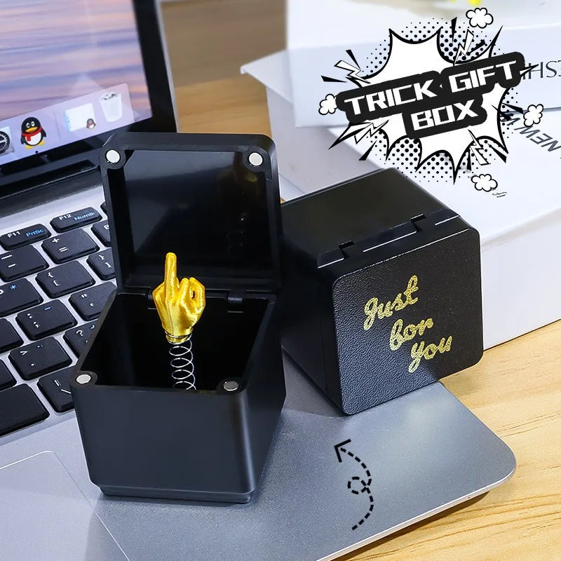 Black box with gold middle finger toy on a desk next to a laptop