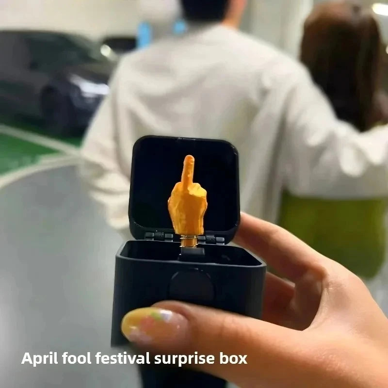 Hand holding a black box with a gold middle finger figurine, labeled 'April fool festival surprise box'.