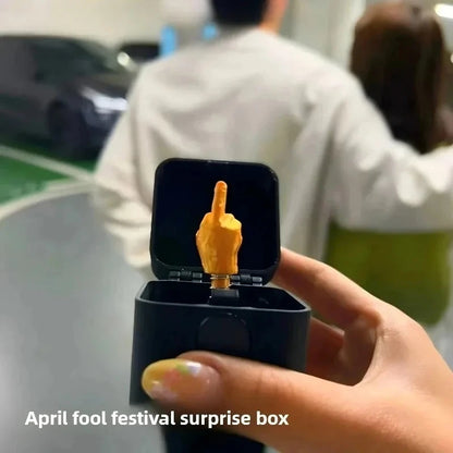 Hand holding a black box with a gold middle finger figurine, labeled 'April fool festival surprise box'.